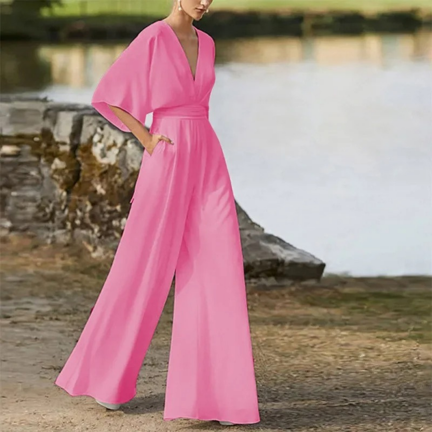 Frau in elegantem, pinkem Jumpsuit mit weitem Bein und tiefem V-Ausschnitt, steht draußen vor einem See. Modetrend, stilvolle Damenbekleidung.