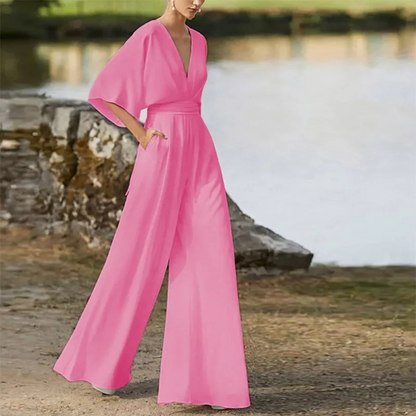 Frau in elegantem, pinkem Jumpsuit mit weitem Bein und tiefem V-Ausschnitt, steht draußen vor einem See. Modetrend, stilvolle Damenbekleidung.
