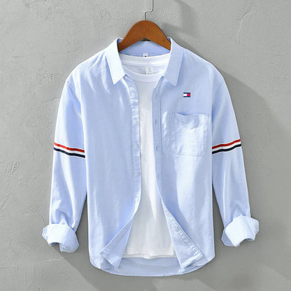 Hellblaues Herrenhemd mit Brusttasche und gestreiften Ärmeln, auf Holzkleiderbügel. Modisches Freizeitkleidungsstück für Männer.