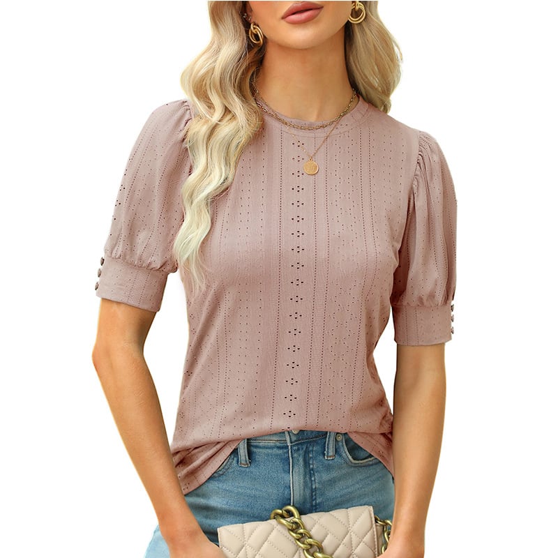 Frau in rosa Spitzenbluse mit kurzen Ärmeln und blonden Haaren, trägt eine goldene Halskette und hält eine beige gesteppte Handtasche.