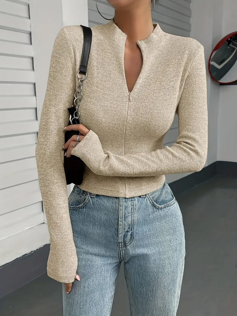 Frau in beigem Strickpullover und blauer Jeans, modisch gekleidet, steht vor einer weißen Wand. Trendige Damenmode, lässiger Look.
