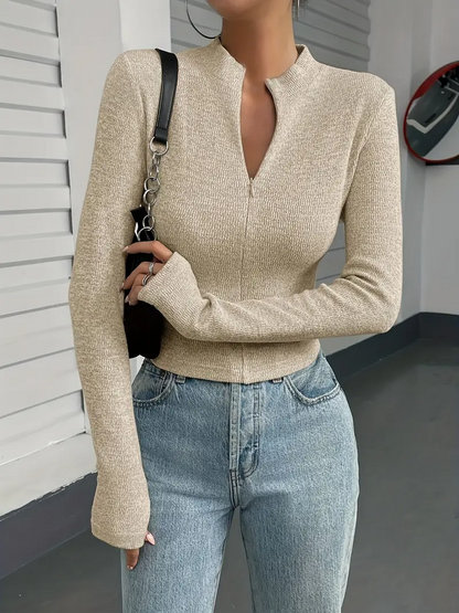 Frau in beigem Strickpullover und blauer Jeans, modisch gekleidet, steht vor einer weißen Wand. Trendige Damenmode, lässiger Look.