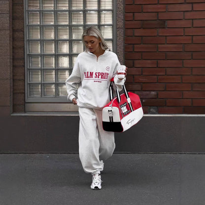 Frau in grauem Jogginganzug mit "Palm Springs"-Aufdruck, trägt rot-weiße Sporttasche, geht vor Backsteinwand. Streetwear, lässiger Stil, urbaner Look.
