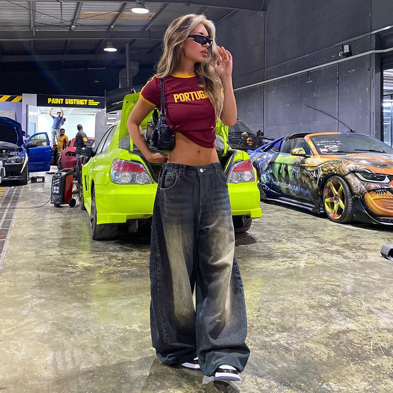 Frau in rotem Portugal-Shirt und weiten Jeans posiert in Autowerkstatt, umgeben von getunten Autos. Urbaner Stil, modische Sonnenbrille.