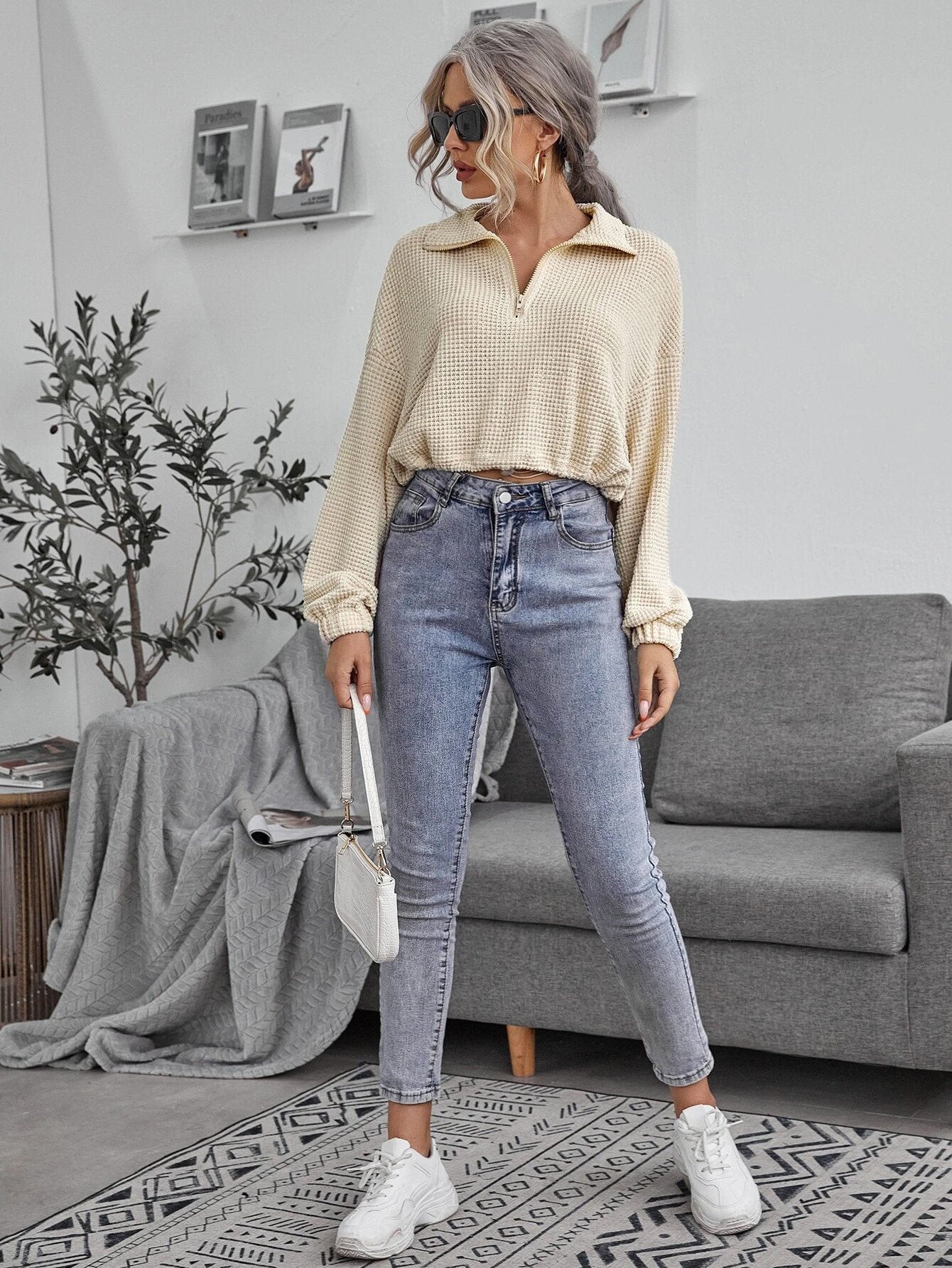 Frau in beiger Bluse und blauer Jeans posiert im modernen Wohnzimmer. Stilvolle Mode, lässiger Look, graues Sofa, Pflanzen, Wanddekoration.
