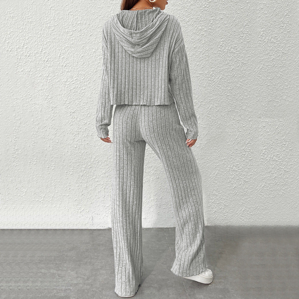 Frau in grauem, geripptem Strick-Zweiteiler mit Kapuze, bestehend aus Pullover und Hose, vor weißer Wand. Bequeme Damenmode, Freizeitlook.