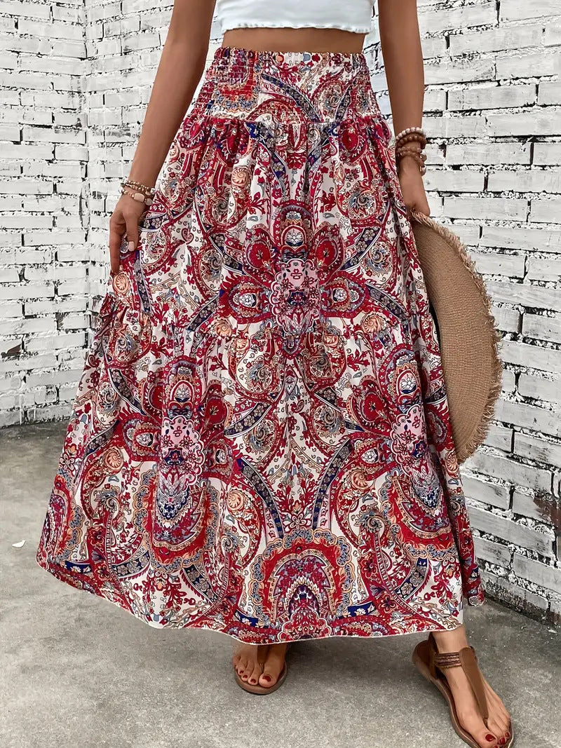 Frau in langem, rotem Boho-Rock mit Paisley-Muster, weißem Top und Sandalen vor weißer Backsteinmauer. Sommermode, Boho-Chic, Damenbekleidung.