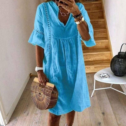 Frau in blauem Sommerkleid mit V-Ausschnitt, hält eine Bambus-Handtasche. Steht in modernem Innenraum mit Holztreppe. Mode, Sommerkleid, Accessoires.