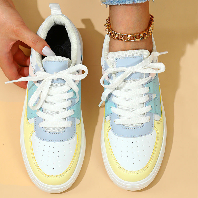Bunte Damen-Sneaker mit weißer Sohle, pastellblauen und gelben Akzenten, geschnürt, getragen mit goldener Fußkette auf beigem Hintergrund.