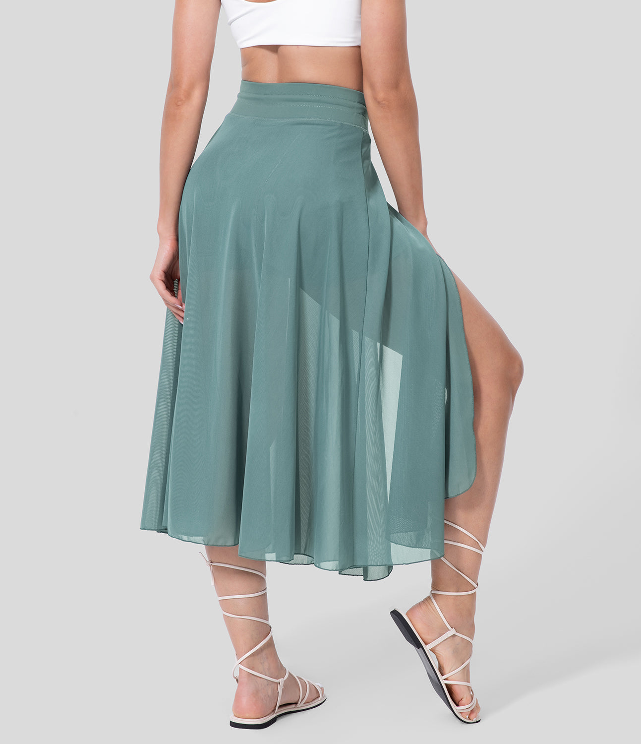 Frau in mintgrünem, transparentem Maxirock mit hohem Schlitz, weißem Crop-Top und Sandalen. Sommermode, eleganter Look, Damenbekleidung.