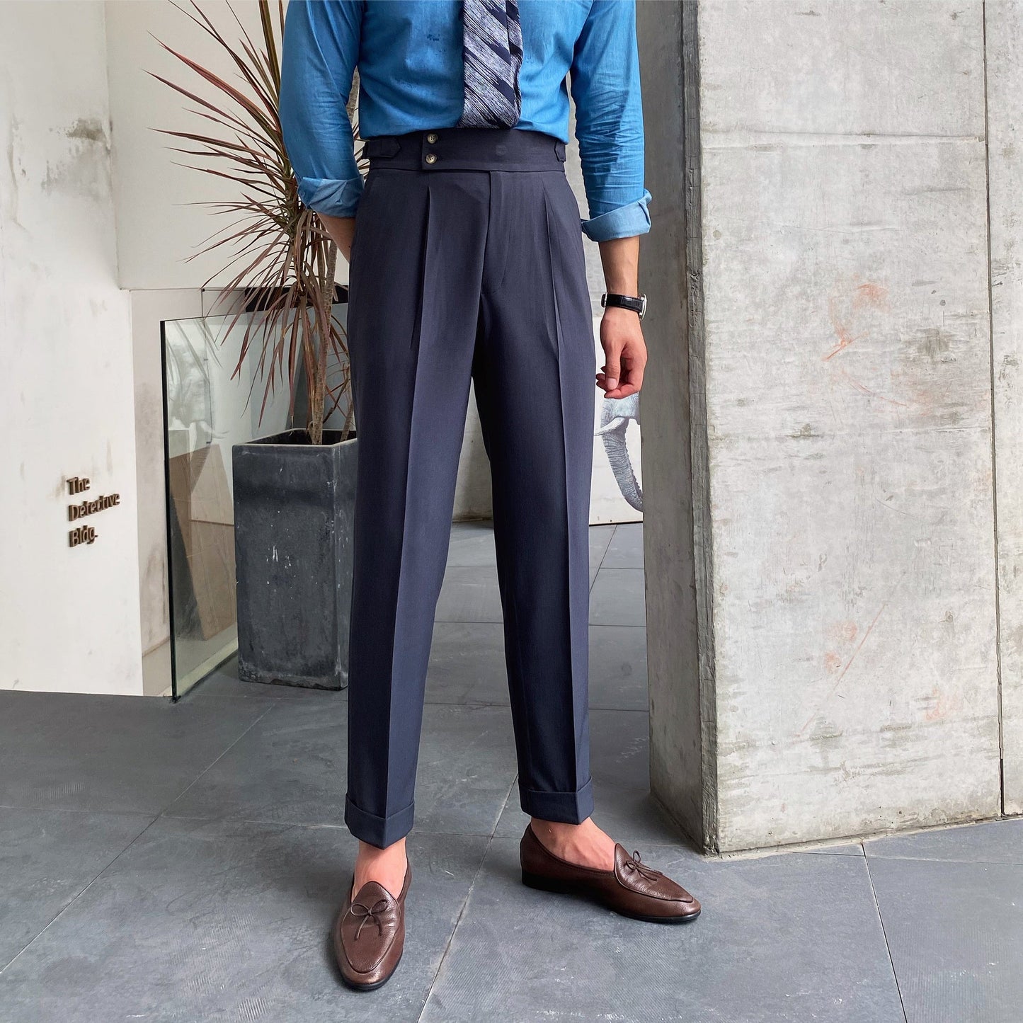 Eleganter Mann in blauer Hemd und Krawatte, trägt dunkelblaue Anzughose und braune Loafer. Steht in modernem Innenraum mit Betonwand und Pflanze.