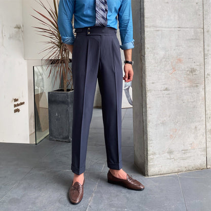 Eleganter Mann in blauer Hemd und Krawatte, trägt dunkelblaue Anzughose und braune Loafer. Steht in modernem Innenraum mit Betonwand und Pflanze.