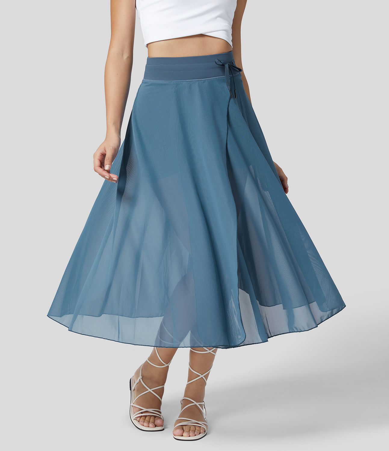 Frau in blauem Chiffon-Wickelrock und weißem Top, modisch, elegant, Sommermode, knöchellang, mit Sandalen, grauer Hintergrund.