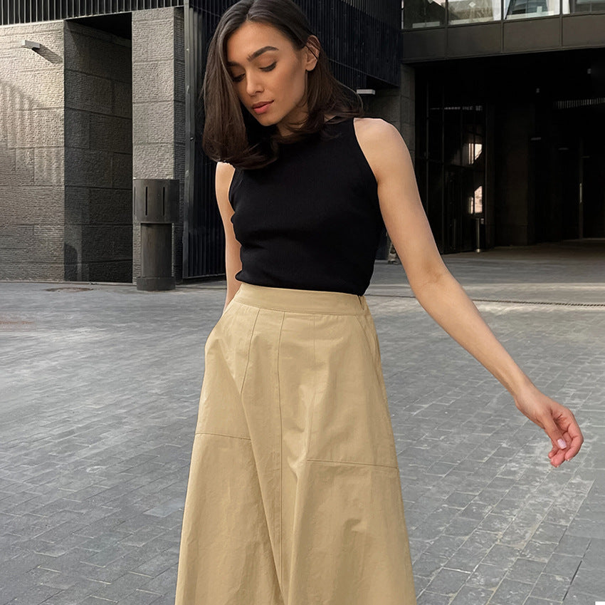 Frau in schwarzem ärmellosen Top und beiger Hose steht auf modernem Stadtplatz. Modetrend, urbane Kleidung, stilvolle Frau, Sommeroutfit.