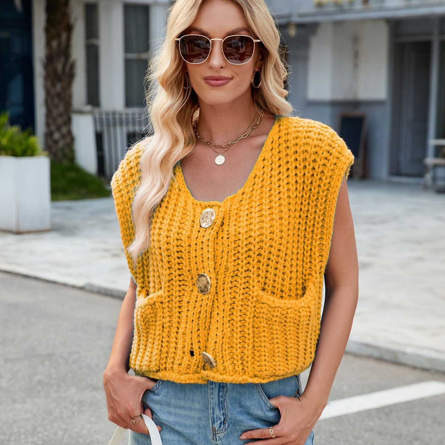 Frau mit blonder Welle, Sonnenbrille, gelber Strickweste mit Knöpfen und Taschen, Jeans, steht draußen. Modetrend, Sommeroutfit, lässiger Stil.