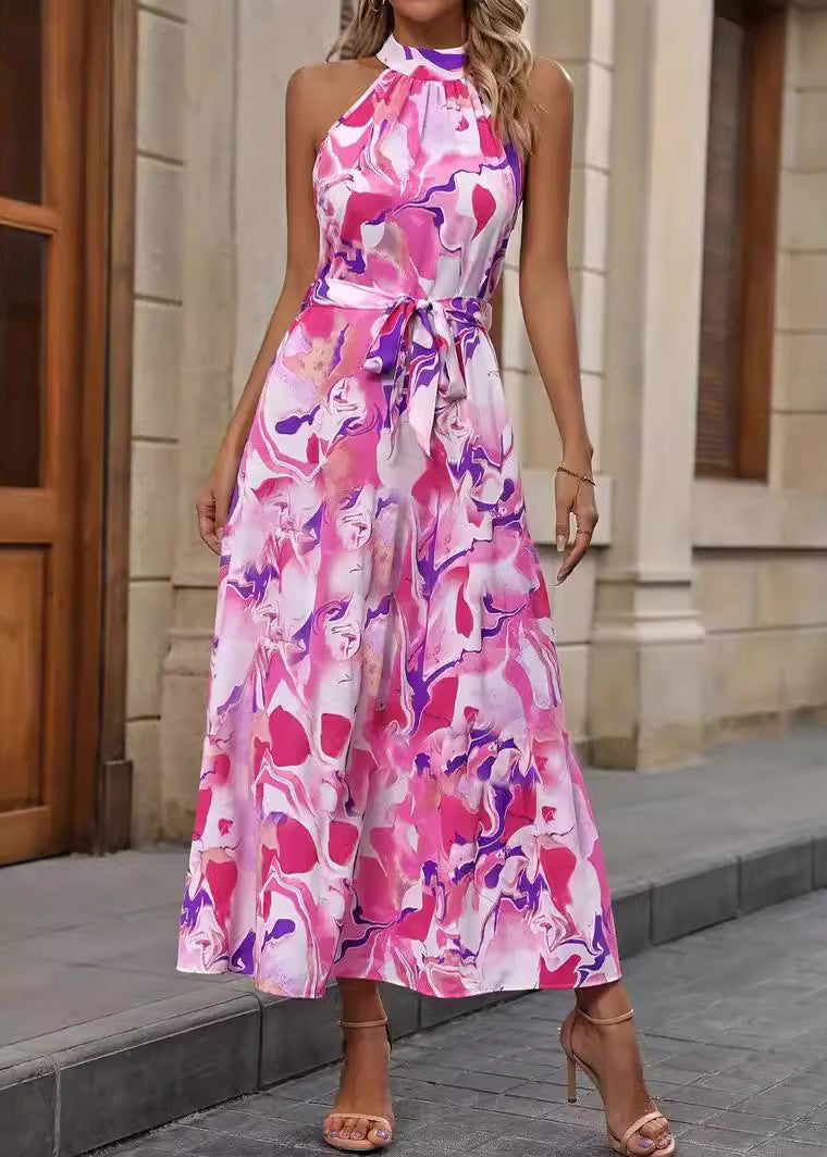 Frau in ärmellosem, pink-weißem Maxikleid mit abstraktem Muster, steht auf Bürgersteig vor Gebäude. Elegante Sommermode, trendiges Design.