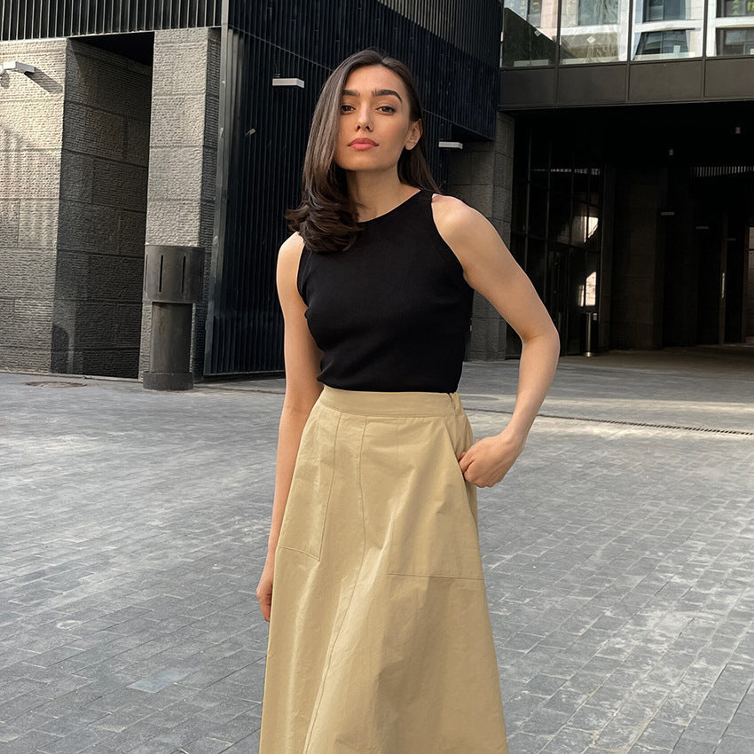 Frau in schwarzem Top und beigem Rock steht auf städtischer Straße. Moderne Architektur im Hintergrund. Mode, Streetstyle, urbane Eleganz.