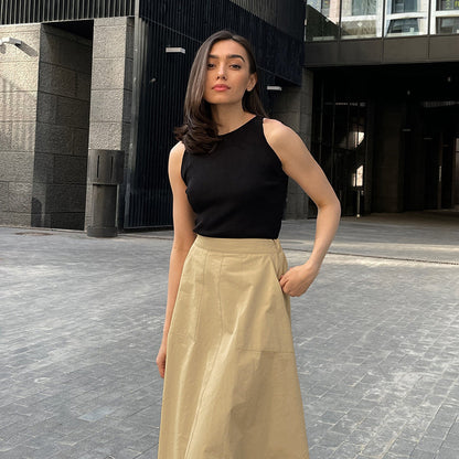 Frau in schwarzem Top und beigem Rock steht auf städtischer Straße. Moderne Architektur im Hintergrund. Mode, Streetstyle, urbane Eleganz.