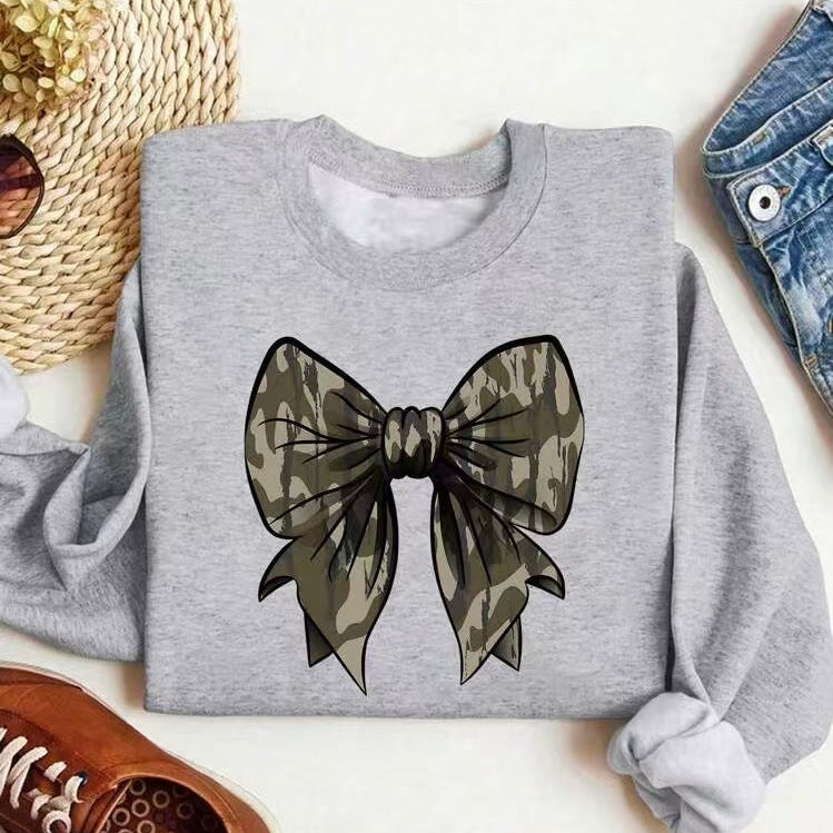 Graues Sweatshirt mit großem Camouflage-Schleifenmotiv, umgeben von Strohhut, Sonnenbrille, Jeans und braunen Schuhen auf weißem Hintergrund.