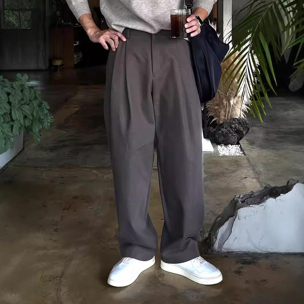 Mann in grauer Hose und weißen Sneakers hält ein Getränk, steht in modernem Innenraum mit Pflanzen. Trendige Herrenmode, lässiger Stil.