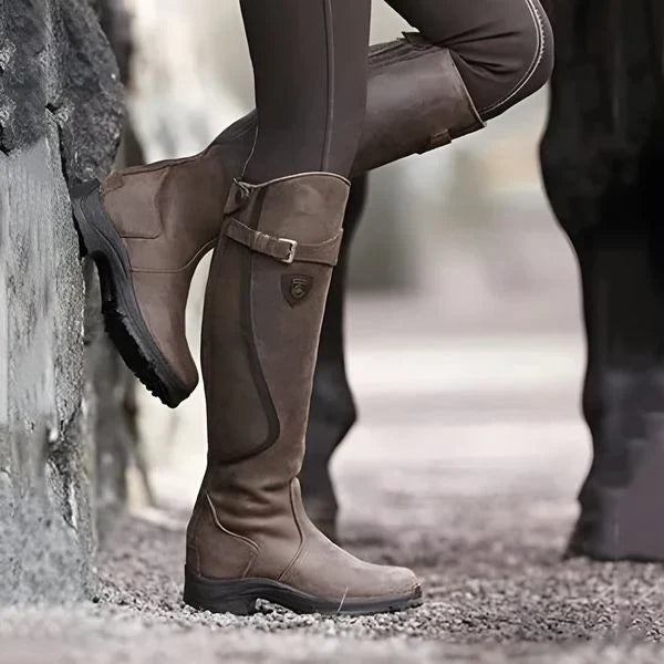Braune Reitstiefel aus Leder, getragen von einer Person in brauner Reithose, lehnt an einer Steinmauer. Outdoor-Reitbekleidung, robustes Design.