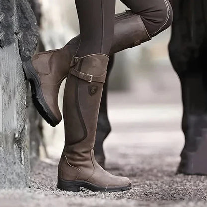 Braune Reitstiefel aus Leder, getragen von einer Person in brauner Reithose, lehnt an einer Steinmauer. Outdoor-Reitbekleidung, robustes Design.