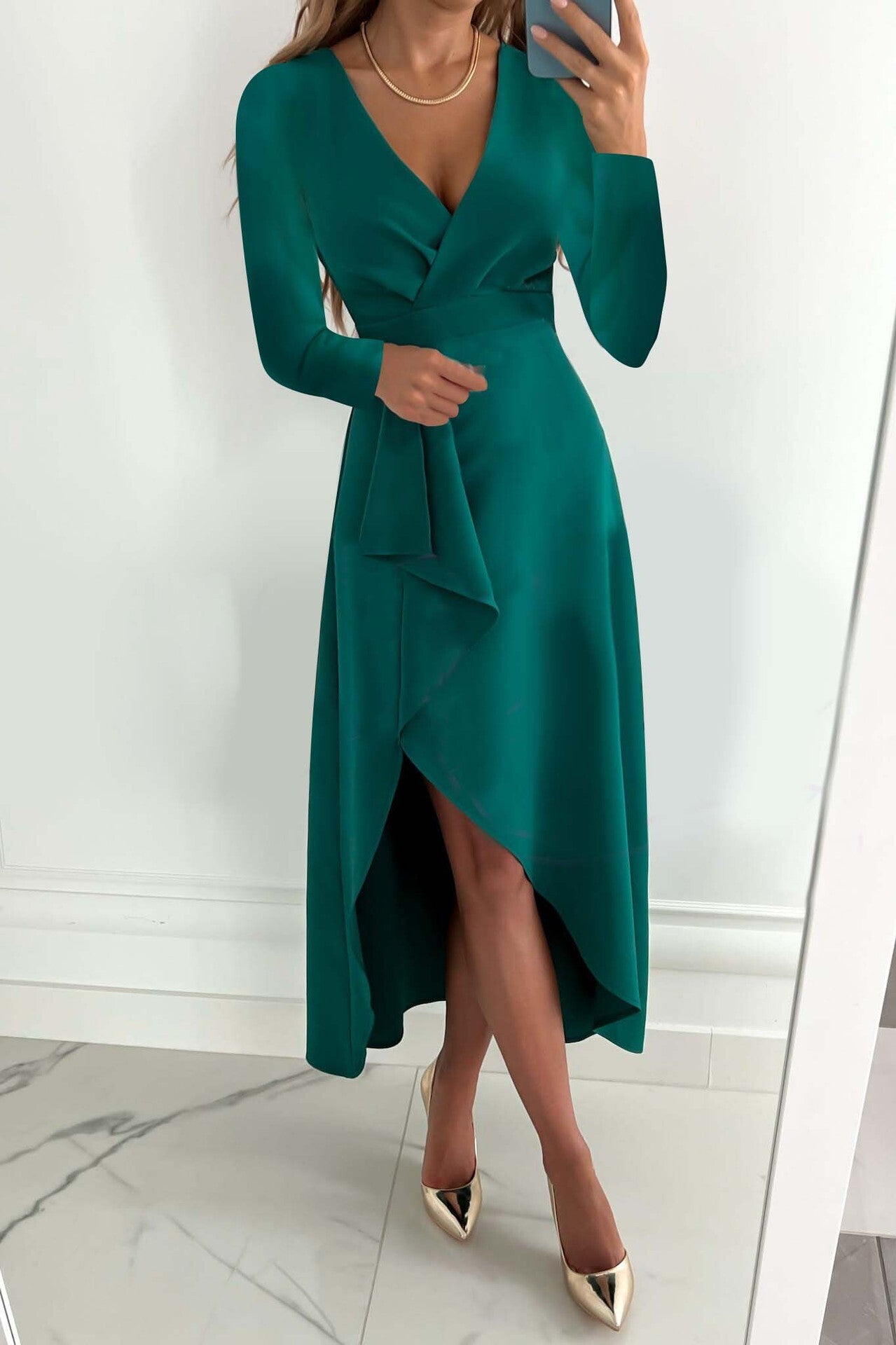 Frau in elegantem, smaragdgrünem Abendkleid mit asymmetrischem Saum und langen Ärmeln, goldene High Heels, Spiegel-Selfie.