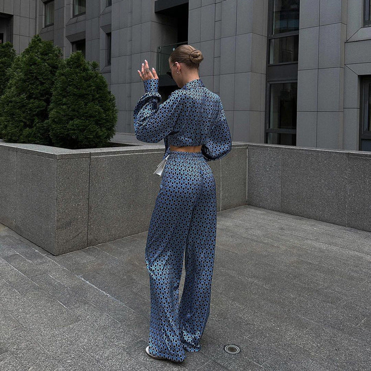 Frau in blauem gemustertem Anzug, steht auf grauem Pflaster vor modernem Gebäude, hebt Hand. Elegante Mode, urbane Architektur, Streetstyle.