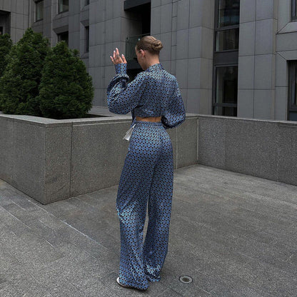 Frau in blauem gemustertem Anzug, steht auf grauem Pflaster vor modernem Gebäude, hebt Hand. Elegante Mode, urbane Architektur, Streetstyle.