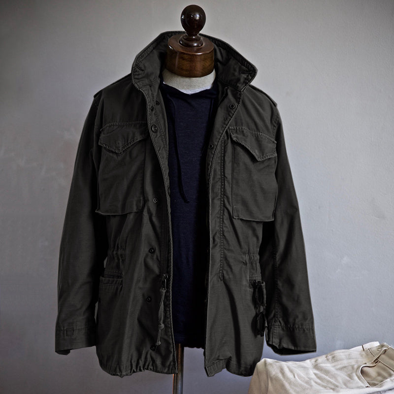 Dunkelgrüne Herren-Feldjacke auf Schneiderpuppe, mit vier Taschen und Kordelzug, über dunkelblauem Hoodie. Modische Outdoor-Bekleidung.