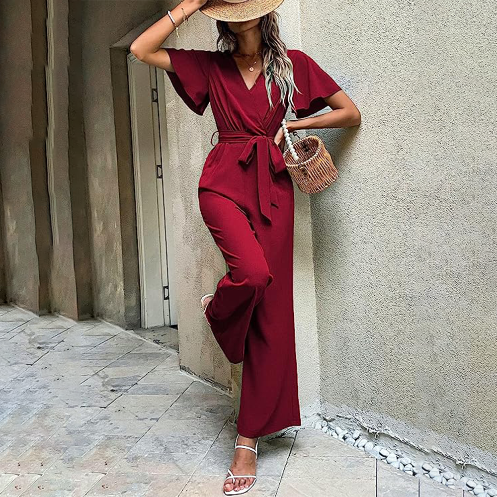 Frau in rotem Jumpsuit lehnt an Wand, trägt Strohhut und weiße Sandalen, hält Korbtasche. Sommermode, eleganter Look, urbane Kulisse.