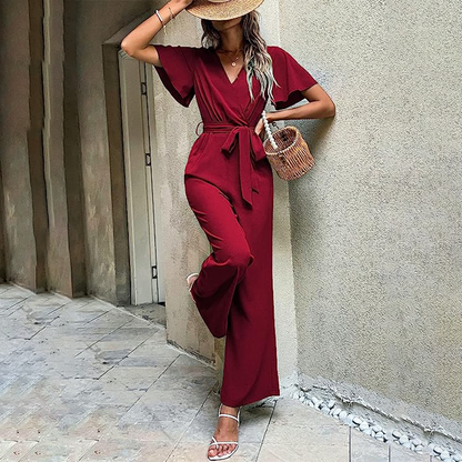Frau in rotem Jumpsuit lehnt an Wand, trägt Strohhut und weiße Sandalen, hält Korbtasche. Sommermode, eleganter Look, urbane Kulisse.