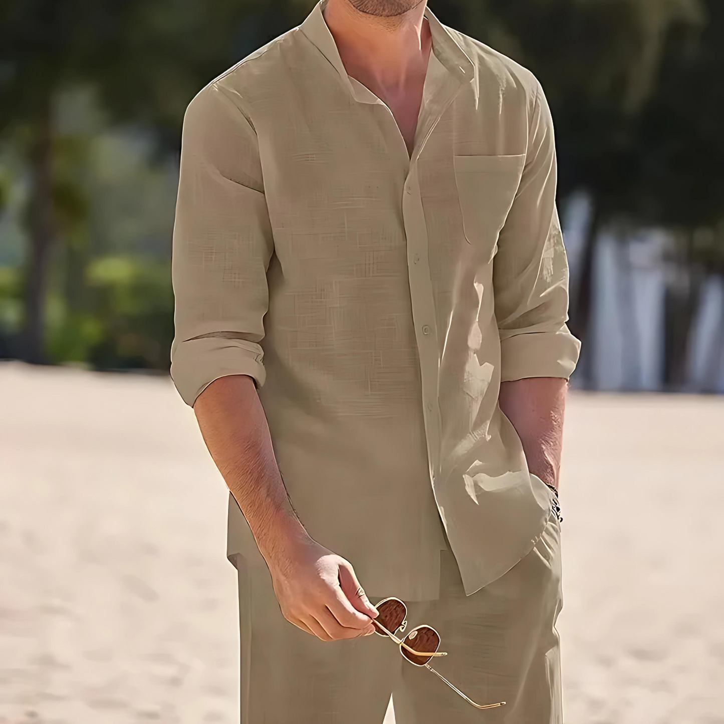 Mann in beigem Leinenhemd und Hose am Strand, hält Sonnenbrille. Sommermode, Freizeitkleidung, stilvoll, entspannt, Strandkulisse.