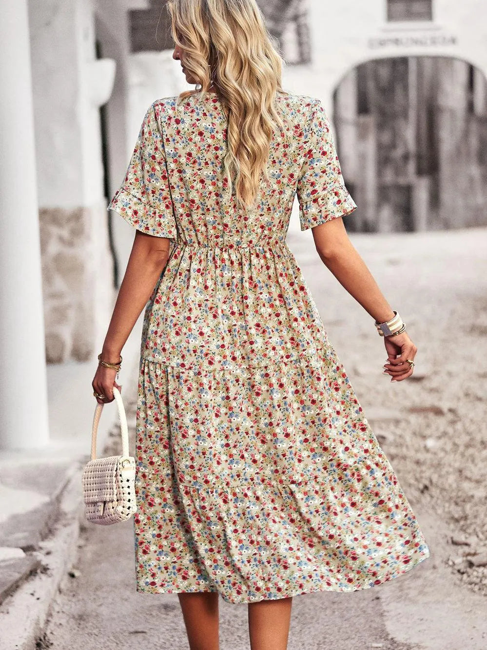 Blonde Frau in geblümtem Sommerkleid, trägt weiße Handtasche, spaziert auf gepflasterter Straße. Modetrend, Damenmode, Sommeroutfit.