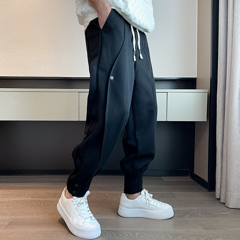 Schwarze Jogginghose mit weißen Kordeln und Knöpfen, kombiniert mit weißen Sneakern auf Holzboden. Moderne Herrenmode, sportlich-eleganter Stil.