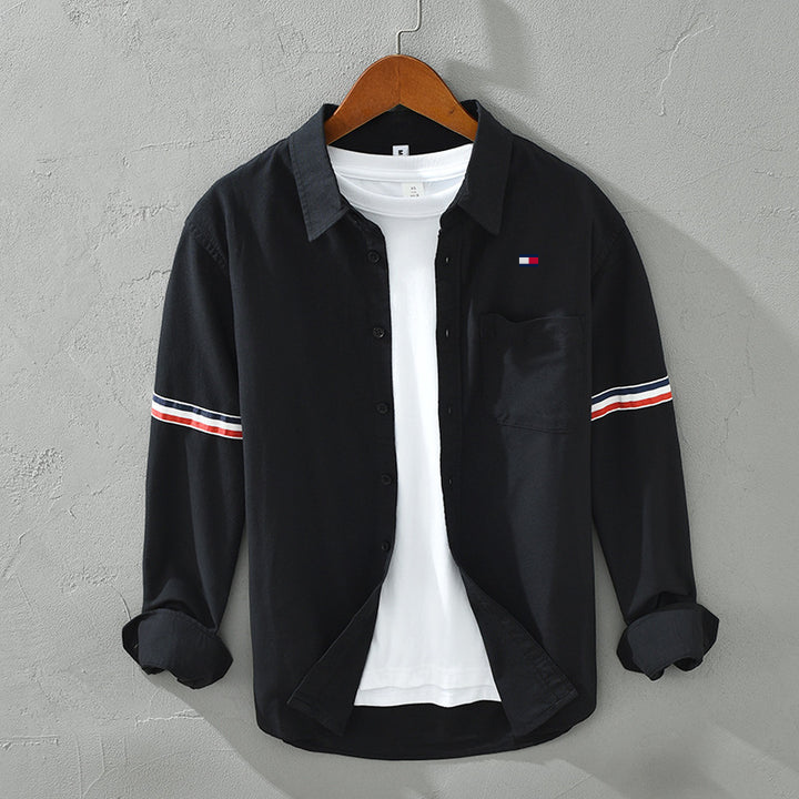 Schwarzes Herrenhemd mit weißen und roten Streifen auf den Ärmeln, über weißem T-Shirt, auf Holzbügel vor grauer Wand. Modetrend, Herrenbekleidung.