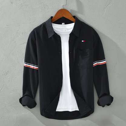 Schwarzes Herrenhemd mit weißen und roten Streifen auf den Ärmeln, über weißem T-Shirt, auf Holzbügel vor grauer Wand. Modetrend, Herrenbekleidung.