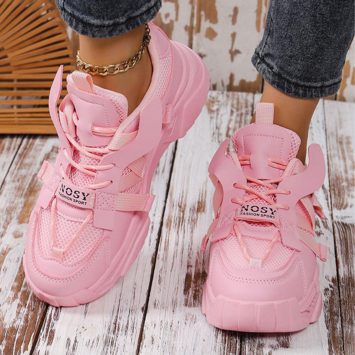 Damen tragen rosa Sneaker mit dicker Sohle und modischem Design, kombiniert mit Jeans und goldener Knöchelkette auf Holzboden.