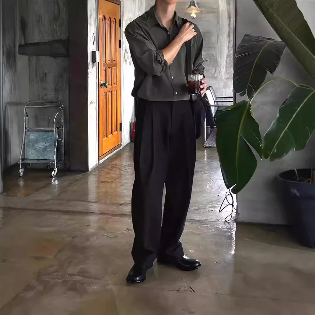 Ein Mann in eleganter, dunkler Kleidung steht in einem modernen Raum mit Pflanzen, hält ein Getränk. Stilvolle Mode, urbaner Lifestyle.