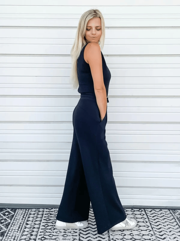 Blonde Frau in schwarzem ärmellosen Jumpsuit und weißen Sneakers vor weißer Wand. Modetrend, lässiger Stil, Sommeroutfit.