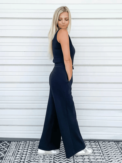 Blonde Frau in schwarzem ärmellosen Jumpsuit und weißen Sneakers vor weißer Wand. Modetrend, lässiger Stil, Sommeroutfit.