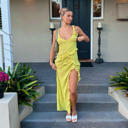 Frau in gelbem Kleid mit Rüschen und hohem Schlitz steht auf Treppe vor Haustür, umgeben von Pflanzen. Elegante Abendmode, Sommerlook.