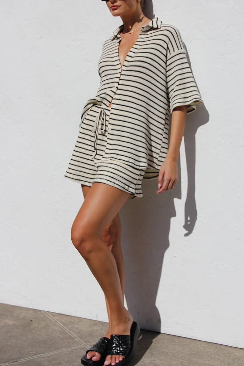 Frau in gestreiftem Sommerkleid vor weißer Wand, trägt schwarze Sandalen. Modetrend 2023, lässiger Look, ideal für warme Tage.