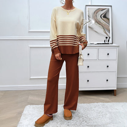 Frau in modischem Herbst-Outfit mit gestreiftem Pullover und brauner Hose, steht vor einer weißen Kommode. Elegante Herbstmode, gemütlicher Look.