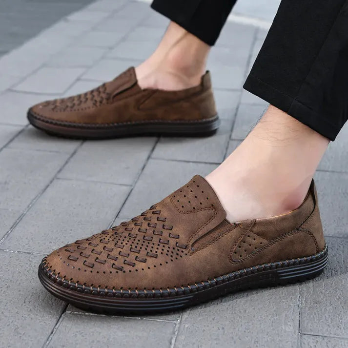 Braune Herren-Slipper aus Leder mit Lochmuster, bequem und atmungsaktiv, auf grauem Pflasterstein getragen. Ideal für Freizeit und Sommermode.