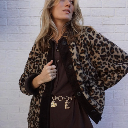 Frau in Leopardenmuster-Jacke vor weißer Ziegelwand, trägt braunes Kleid und goldene Gürtelkette. Modetrend, Herbstmode, Streetstyle.