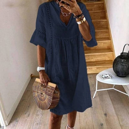 Frau in dunkelblauem Sommerkleid mit V-Ausschnitt, hält eine Bambus-Handtasche. Steht in modernem Flur mit Holztreppe und Dekoration.
