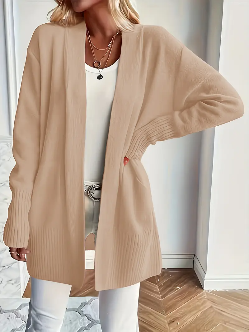 Frau trägt beige Strickjacke, weißes Top und weiße Hose. Modischer Herbst-Look, lässiger Stil, elegantes Outfit, Damenmode, Indoor-Aufnahme.
