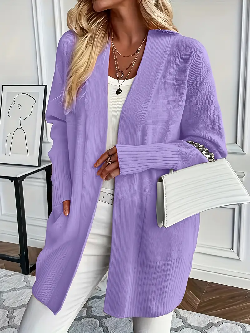Frau in lila Strickjacke, weißem Top und Hose, hält weiße Handtasche. Moderne, stilvolle Mode vor minimalistischem Hintergrund.