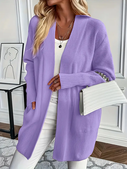 Frau in lila Strickjacke, weißem Top und Hose, hält weiße Handtasche. Moderne, stilvolle Mode vor minimalistischem Hintergrund.